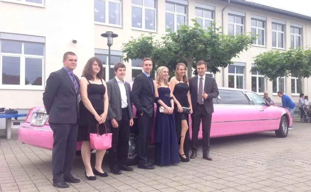 Junge Leute in festlicher Kleidung stehen vor der pinken Stretchlimousine vor einer Schule