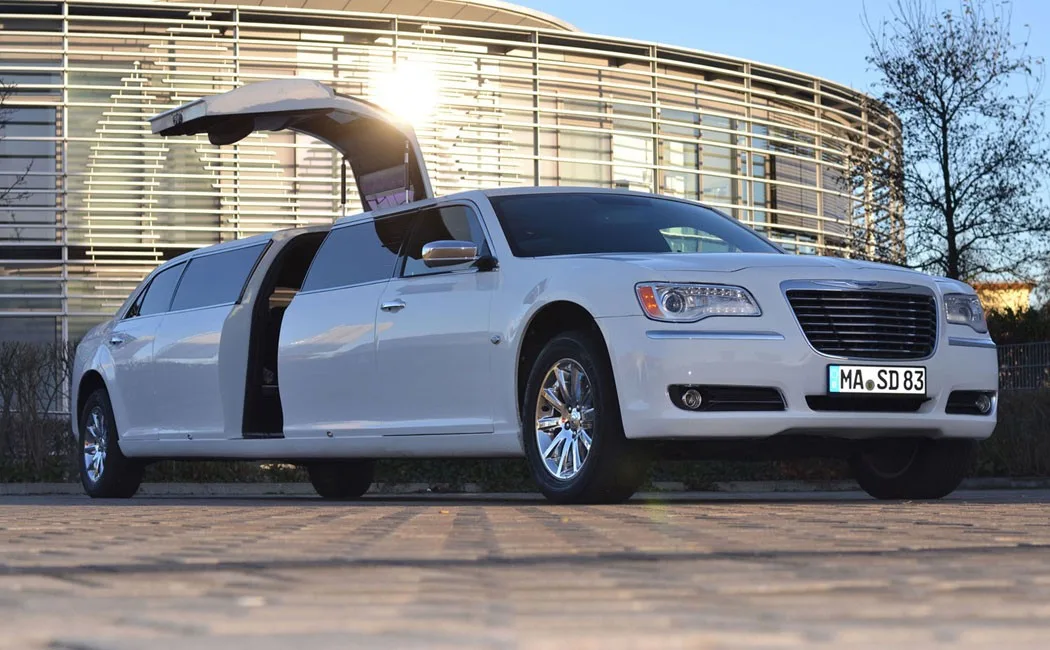 Weißer Chrysler 300C mit geöffneter Flügeltür vor einem modernen Gebäude im Abendlicht