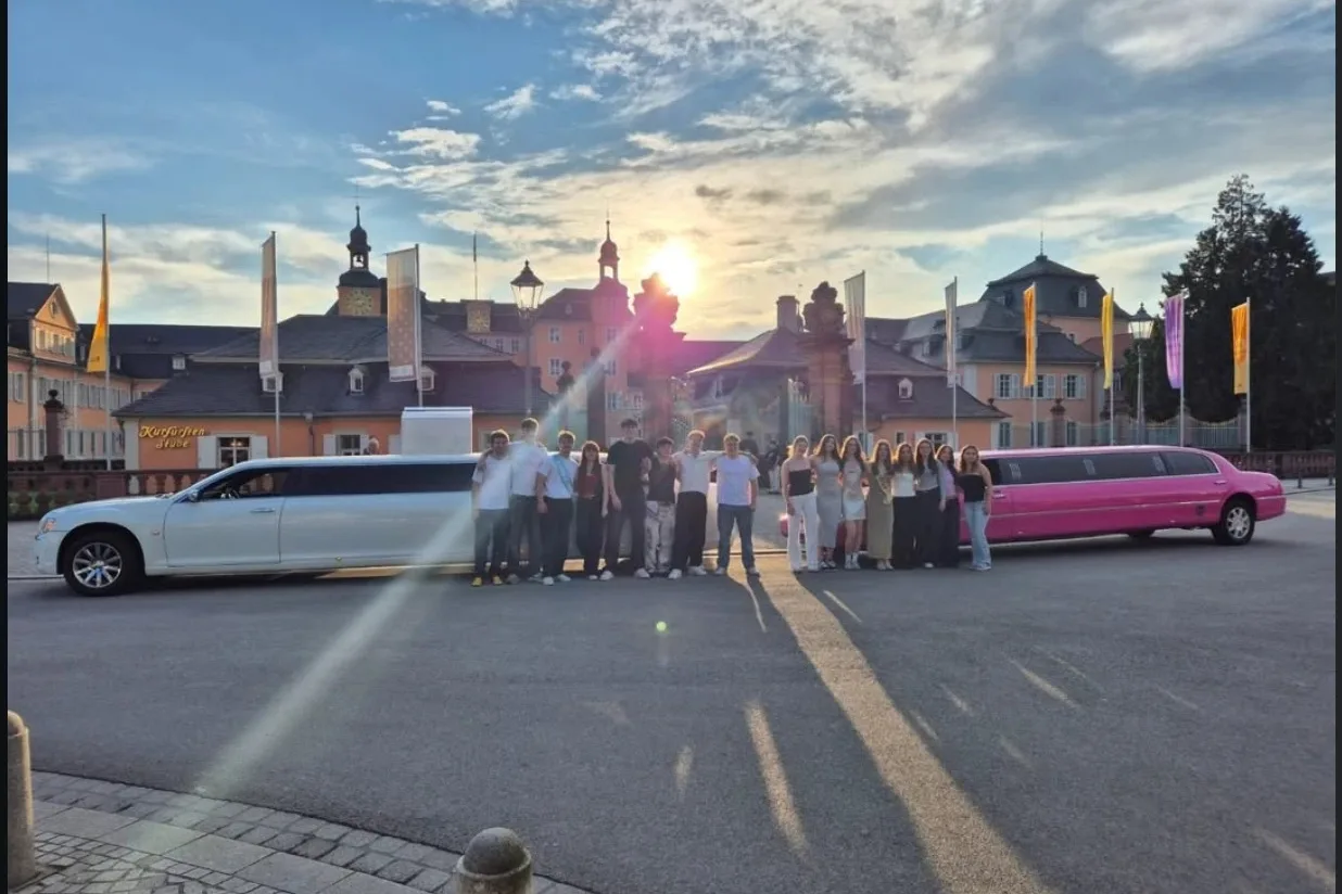 Gruppe junger Leute steht zwischen einer weißen und einer pinken Stretchlimousine vor einem Schloss im Sonnenuntergang