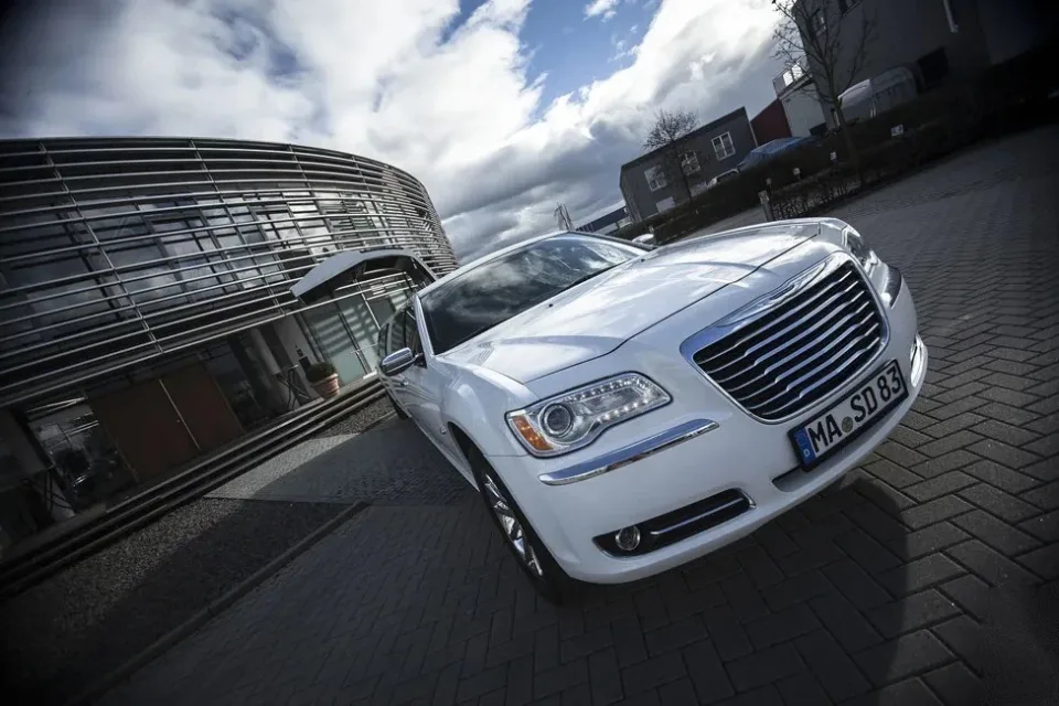 Weißer Chrysler 300C von vorn vor einem modernen Gebäude mit Wolkenhimmel